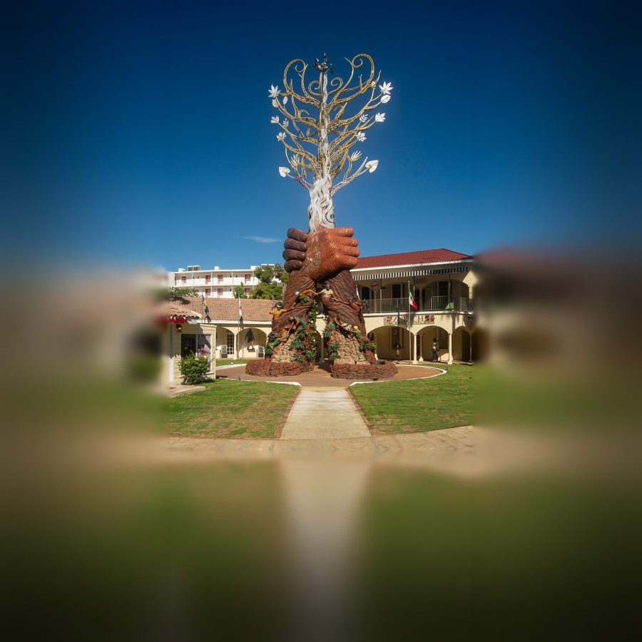 Together We Rise: two vast clasped hands carved like tree trunks rise from a rock base, holding up a gold and silver Tree of Life against a blue sky